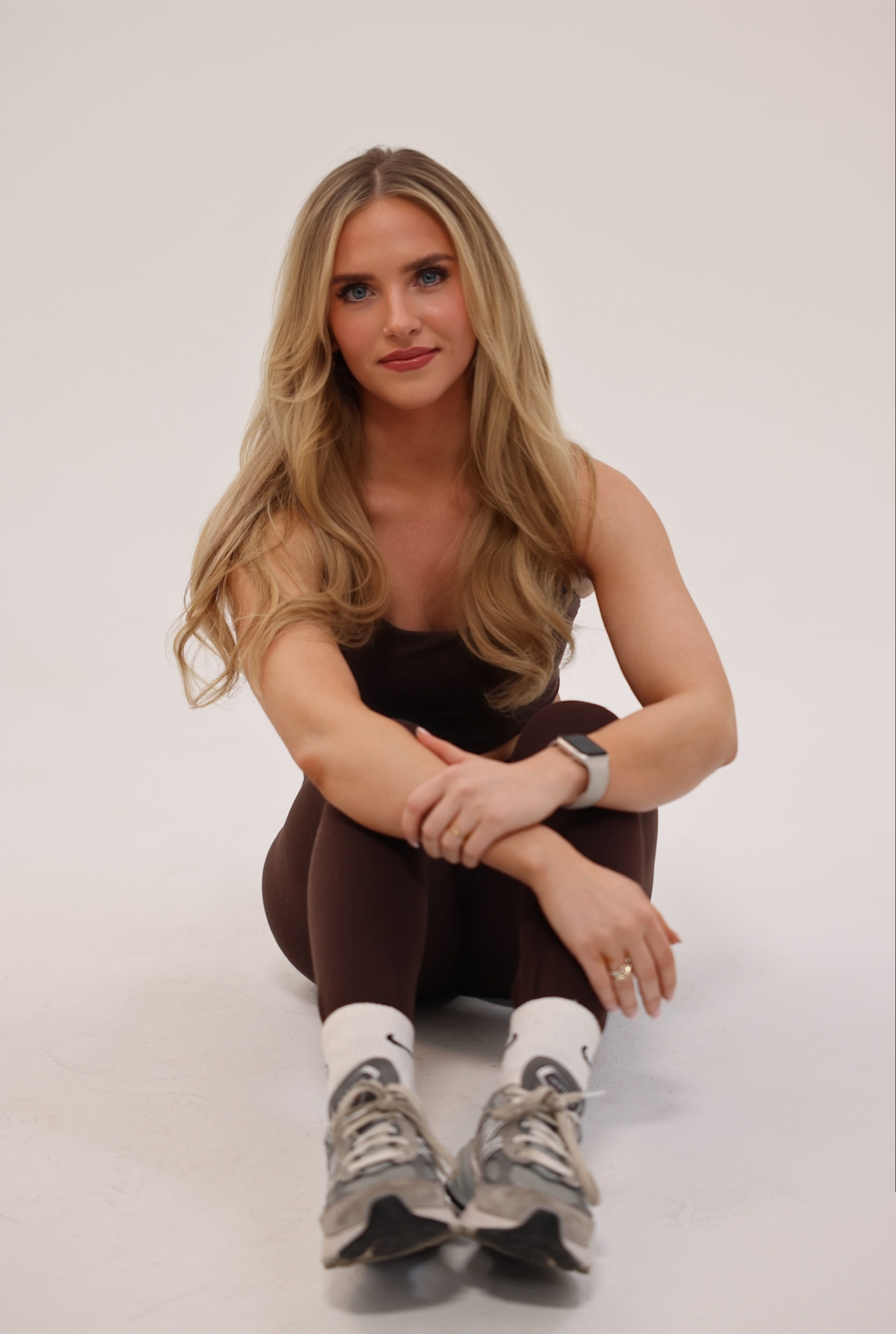 Holland Scattergood, a blonde woman in a fitness outfit is sitting on the ground with her hands clasped looking into the camera.